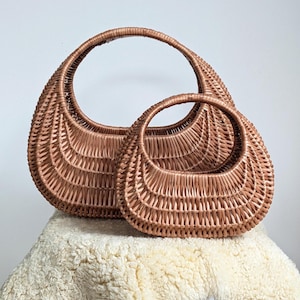May include: Two woven rattan handbags in a warm brown hue. The bags have a rounded, crescent shape with a sturdy handle. The larger bag is approximately 14 inches wide, and the smaller bag is about 10 inches wide. The bags are displayed on a fluffy, off-white surface.