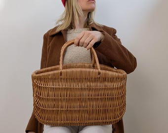 Puede incluir: Una cesta rectangular de mimbre trenzado con un acabado natural en color marrón claro, que presenta un diseño robusto de parte superior abierta y dos asas arqueadas unidas al borde superior, tejida con tiras de caña finas y flexibles.