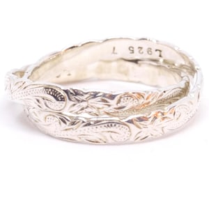 May include: A silver ring with an intricate floral pattern. The ring is made of intertwined bands, with the text "L925 T" visible on the inner band. The ring has a polished finish and is set against a white background.