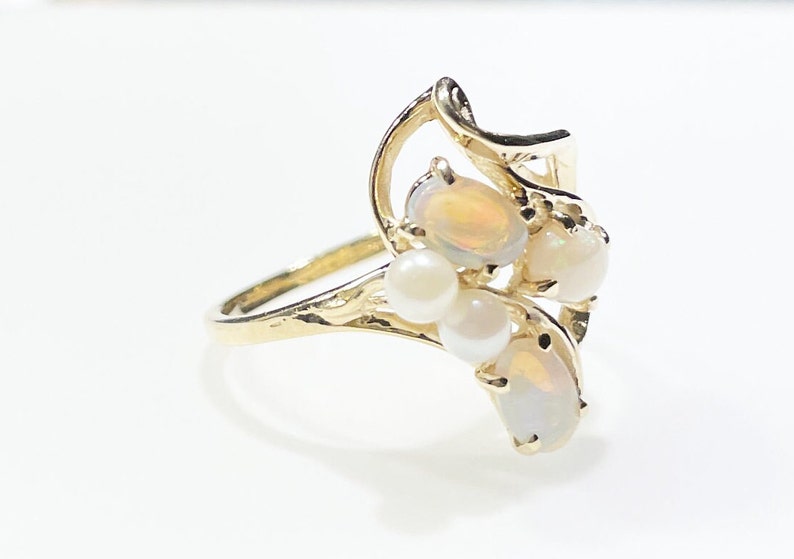 Pu&ograve; includere: Un anello d'oro con un gruppo di opali e perle. L'anello ha un design delicato e a spirale.