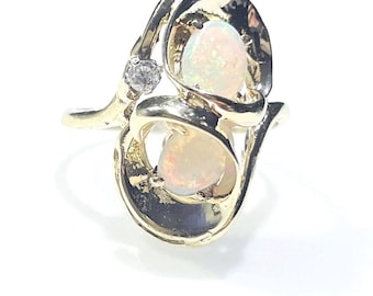 May include: A gold ring featuring two oval-shaped opals with iridescent flashes of color, set within a swirling, organic metal frame. A single, small, clear round-cut gemstone is positioned on the side of the band, accenting the design.