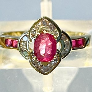 May include: A gold ring with an oval ruby centre stone, encircled by diamonds. The band includes rectangular rubies, enhancing the ring's classic design. The ring is displayed against a neutral backdrop, highlighting its features.