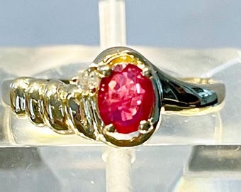 May include: A gold ring with a red oval gemstone and a smaller diamond accent. The ring band is twisted and has a textured design.