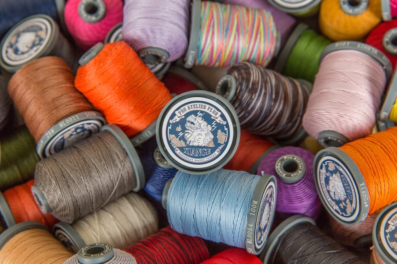 May include: A close-up of a variety of spools of thread in different colors, including orange, pink, blue, and green. The spools are labeled with the text "Pro Atelier Plus" and "NIANCE Handmade Waxed Thread".