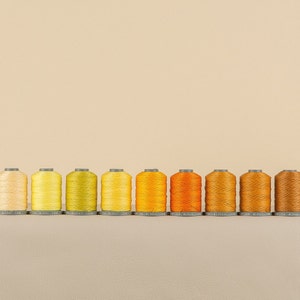 May include: Ten spools of thread in various shades of yellow, orange, and brown. The spools are arranged in a row from lightest to darkest.