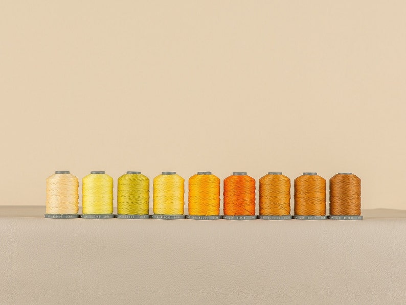 May include: Ten spools of thread in various shades of yellow, orange, and brown. The spools are arranged in a row from lightest to darkest.