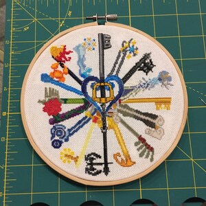 May include: Cross-stitch embroidery art featuring a collection of stylized keys in various colors, arranged in a radial pattern. The keys are centered around a heart-shaped key design. The artwork is displayed in a wooden embroidery hoop.