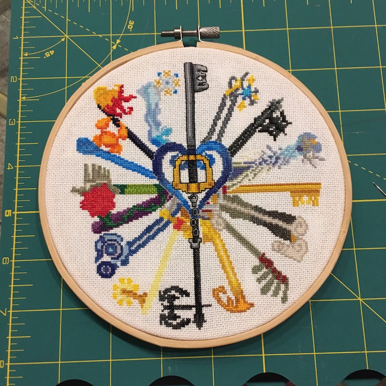 May include: Cross-stitch embroidery art featuring a collection of stylized keys in various colors, arranged in a radial pattern. The keys are centered around a heart-shaped key design. The artwork is displayed in a wooden embroidery hoop.