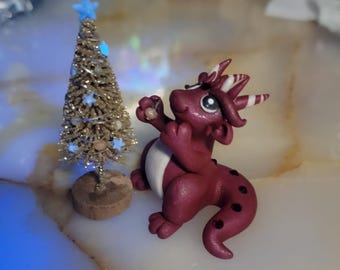 May include: A miniature Christmas tree with a blue star topper and a small, maroon dragon figurine. The dragon has a white belly, dark spots, and red horns. The tree is decorated with small, pearl-like ornaments.