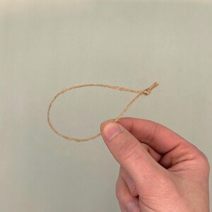May include: A loop of natural-colored twine, knotted at one end. The twine is held by a hand against a light green background. The twine is thin and appears to be made of natural fibers. The loop is approximately 4 inches in diameter.