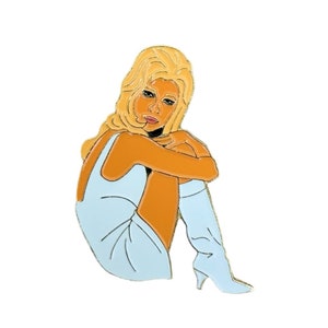 Op de afbeelding: Een gouden emaille pin met een vrouw met blond haar die een lichtblauwe jurk en witte laarzen draagt. Ze zit met haar benen gekruist en haar armen om haar benen geslagen.