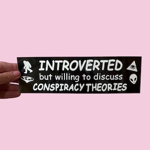 May include: A black sticker with white text that reads "INTROVERTED but willing to discuss CONSPIRACY THEORIES." The sticker features illustrations of a Bigfoot, a UFO, an eye inside a triangle, and an alien head.