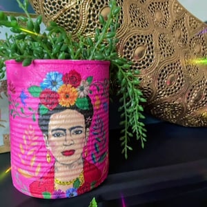 May include: A pink painted tin can with a colorful image of Frida Kahlo, flowers, and foliage. The can is filled with a green succulent plant.