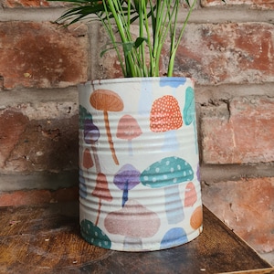 Op de afbeelding: Een witte blikken plantenpot met een kleurrijk paddenstoelenpatroon. De pot is gevuld met een groene, bladerrijke plant.