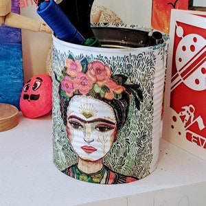 Puede incluir: Una lata de metal blanca con un diseño colorido que presenta el rostro de una mujer con flores en el pelo. La lata se utiliza como porta lápices y contiene tijeras y marcadores.