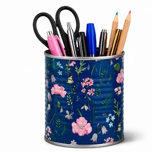 May include: A cylindrical metal pencil holder featuring a deep blue background decorated with a colourful pattern of various pink, white, and light blue wildflowers and green foliage. The container is filled with black scissors, pens, and pencils.