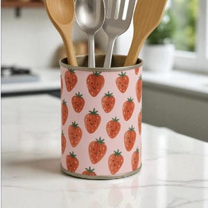 May include: A pink utensil holder decorated with smiling strawberry motifs. It holds a wooden spoon, a metal spoon, a metal spatula, and a wooden spatula. The holder sits on a white countertop.
