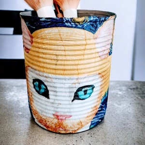 May include: A decorative tin can featuring a cat face design with blue eyes and orange fur. The can holds two makeup brushes with peach-colored bristles. The can is decorated with blue and yellow patterns.