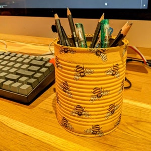 May include: A yellow can with a bee pattern is used as a pencil holder. It is filled with pencils, pens, and markers.