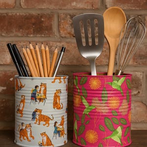 May include: Two decorative tin cans used as utensil holders. One is light grey with a tiger and people design, the other is pink with a green and yellow floral pattern. The left can holds pencils and pens.