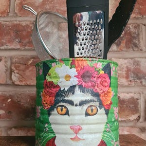 May include: A cylindrical metal kitchen utensil holder featuring a colorful graphic of a cat wearing a floral crown. The container is filled with a silver mesh strainer, a black-handled metal grater, and black kitchen tongs, all standing upright.