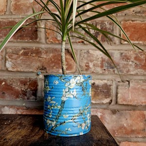 Puede incluir: Una lata de metal pintada de azul reutilizada como maceta, con un diseño floral que recuerda a una pintura famosa. La planta tiene hojas largas y verdes con bordes rosados. El fondo es una pared de ladrillos.