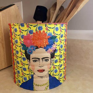 May include: A cylindrical metal container featuring a vibrant yellow background with a repeating blue and red floral pattern. The front displays a detailed illustration of a person with dark hair adorned with colourful orange, pink, and blue flowers.