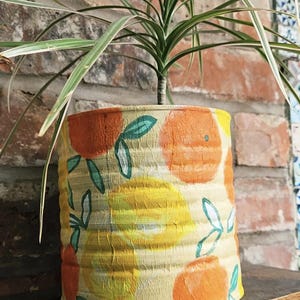 May include: A cylindrical plant pot with a painted design of oranges and leaves. The pot is filled with a green plant. The background is a brick wall.