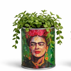 May include: A cylindrical metal planter featuring a colorful portrait of Frida Kahlo with red flowers in her hair, gold earrings, and a gold necklace. The planter contains a green succulent plant with small, rounded leaves spilling over the top edge.