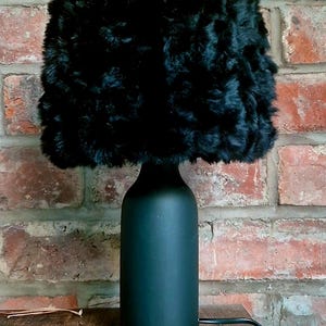 May include: A table lamp featuring a cylindrical matte black base and a textured black faux fur lampshade. The lamp has a smooth, solid-colored base and a fluffy, high-pile shade that provides a distinct contrast in texture and material finish.