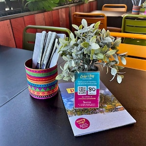 Può includere: Un tavolo nero con una lattina colorata che contiene posate, una piccola pianta verde in un vaso e un cartello blu e bianco con un codice QR e il testo "Order & Pay" e "80". Il cartello è su una brochure piegata con una scena di spiaggia e il testo "Happy Days!"