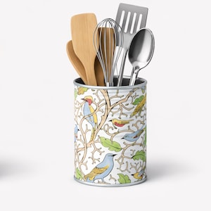 May include: A cylindrical kitchen utensil holder featuring a white background with a repeating pattern of colourful birds, light green leaves, and tan branches. The container holds two wooden spatulas, a metal whisk, a metal spatula, and a metal spoon.