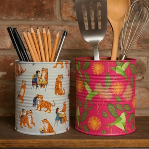 May include: Two decorative tin cans. One features a tiger print and holds pens and pencils. The other, with a floral design, contains a spatula, wooden spoon, and whisk. Both are on a wooden shelf, against a brick wall.