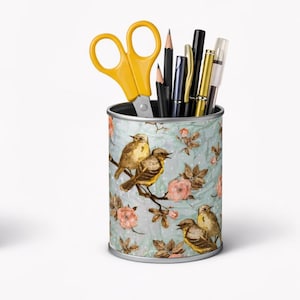 May include: A cylindrical metal pencil holder featuring a light blue background with a pattern of brown birds perched on branches and pink flowers. The container is filled with yellow scissors, two graphite pencils, and four pens.