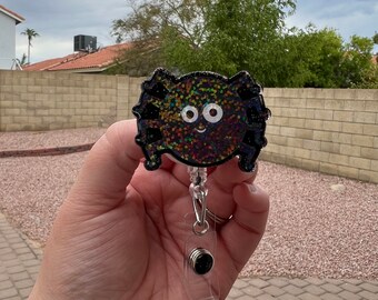May include: A black glitter spider-shaped badge reel with a smiling face and white eyes. The badge reel has a clear plastic holder and a silver clip. The background is a cloudy sky and a brick wall.