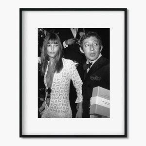 May include: A black and white photograph of a woman wearing a white lace dress and a man wearing a suit and bow tie. The man is holding a box in his right hand.