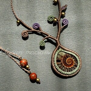 May include: Handmade macrame necklace featuring a fossil pendant with a spiral design. The pendant is surrounded by green and brown macrame, with purple and green spiral accents. The necklace also includes a separate strand with red and gold beads.