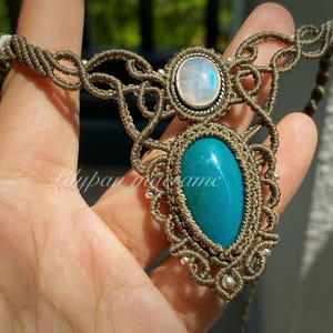May include: Handmade macrame necklace featuring a large, oval turquoise stone and a smaller, round, iridescent stone. The necklace is crafted with brown cord and accented with small silver beads. The design is intricate and detailed, showcasing the craftsmanship.