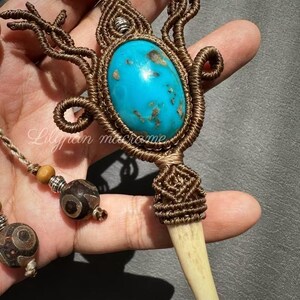 Puede incluir: Un collar de macramé hecho a mano con un gran colgante ovalado de piedra turquesa. El colgante está intrincadamente tejido con cordón marrón e incluye un dije en forma de hueso. Se ven acentos de cuentas adicionales y el texto "Lilyan macrame".