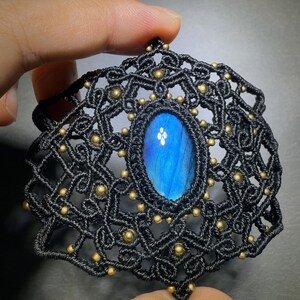 May include: A handcrafted black macramé pendant featuring an oval, iridescent blue gemstone. The intricate woven design is accented with small, gold-coloured beads. The pendant is held up by a hand.