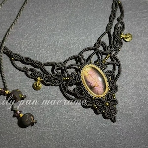 Op de afbeelding: Zwarte macramé ketting met een centrale ovaalvormige steen in roze en goudtinten. De ketting heeft ingewikkeld knoopwerk en kleine goudkleurige accenten. De tekst "Lily pan macrame" is zichtbaar.