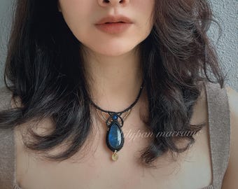 Puede incluir: Un collar de macramé negro con un gran colgante de piedra preciosa azul en forma de lágrima. El collar tiene detalles dorados y un pequeño dije redondo dorado. El texto "lilypan macrame" es visible en el lado derecho de la imagen.
