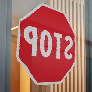 May include: A red octagonal sticker applied to a glass surface, featuring a white border and the word STOP printed in large white capital letters. The text appears reversed as if viewed from the opposite side of the transparent glass pane.