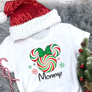 May include: White t-shirt featuring a red and green candy cane Mickey Mouse design with the word "Mommy" printed on it. The shirt is styled with a red and white Santa hat and a small Christmas tree.