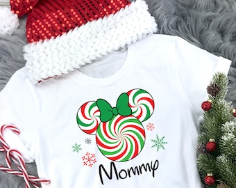 May include: White t-shirt featuring a red and green candy cane Mickey Mouse design with the word "Mommy" printed on it. The shirt is styled with a red and white Santa hat and a small Christmas tree.