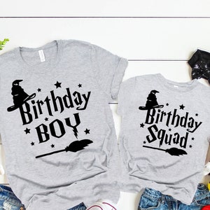 Puede incluir: Dos camisetas grises con texto y gráficos negros. La camiseta más grande dice "Birthday Boy" con un sombrero de bruja y una escoba. La camiseta más pequeña dice "Birthday Squad" con un sombrero de bruja y una escoba.