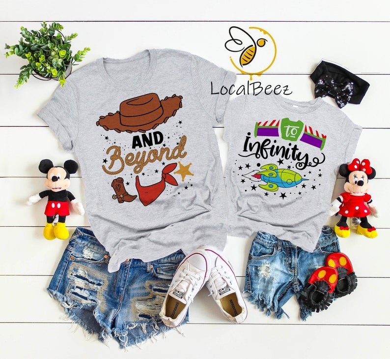 May include: Two gray t-shirts with a Toy Story theme. The larger shirt has a brown cowboy hat, red bandana, and the text "And Beyond". The smaller shirt has a cartoon spaceship and the text "To Infinity".