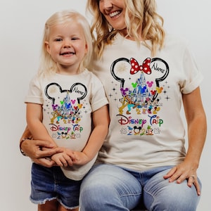 May include: Two white t-shirts with a Mickey Mouse head design. The design features Mickey Mouse's ears, a castle, and other Disney characters. The text "Disney Trip" is printed on the shirts. The shirts are personalized with the text "Name".