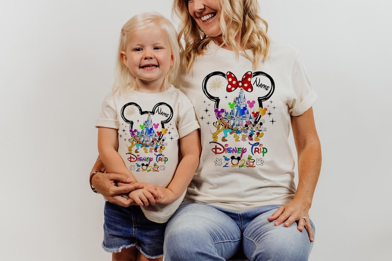 May include: Two white t-shirts with a Mickey Mouse head design. The design features Mickey Mouse's ears, a castle, and other Disney characters. The text "Disney Trip" is printed on the shirts. The shirts are personalized with the text "Name".