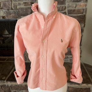 May include: A long-sleeved button-down shirt in a solid peach color featuring a ruffled collar, a front button placket, and a small embroidered logo on the left chest. The shirt is displayed on a mannequin and has cuffed sleeves.